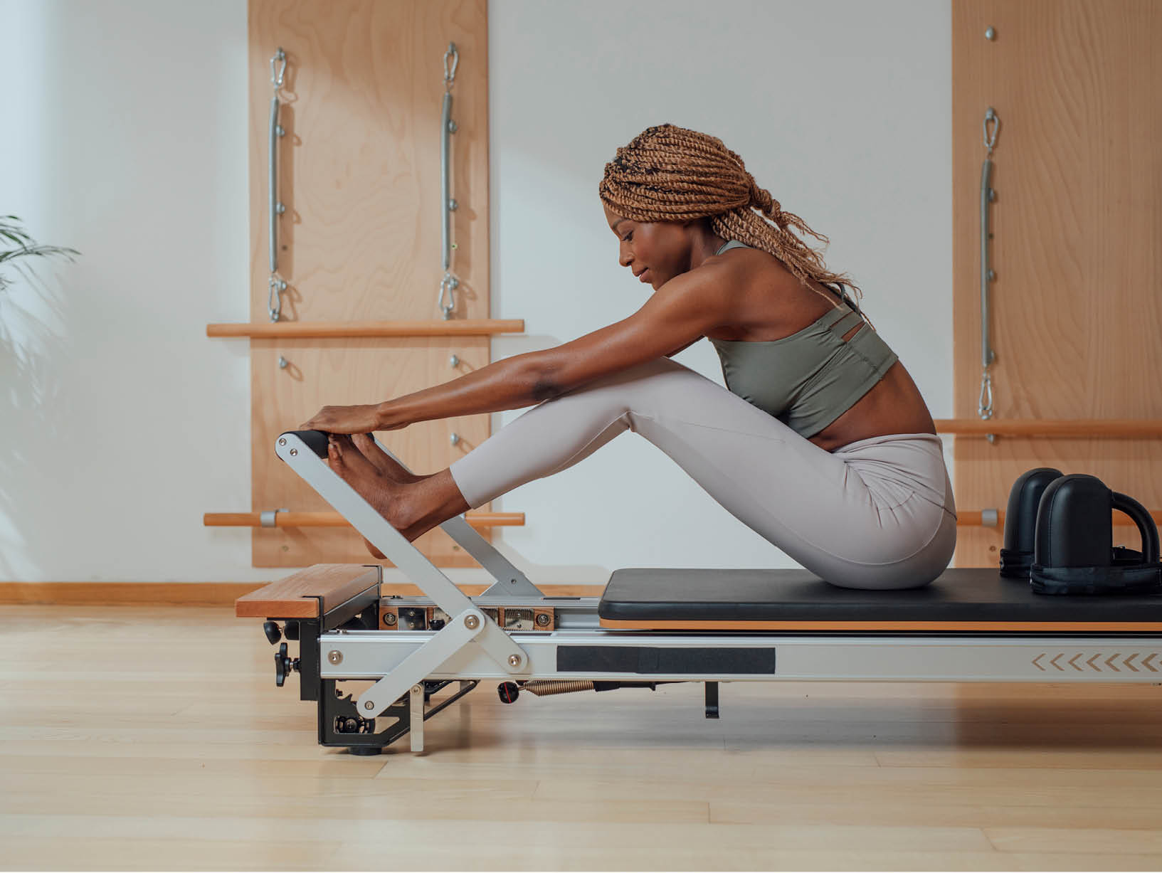 A woman practicing a seated stretch on a Pilates reformer in a bright, sunlit studio